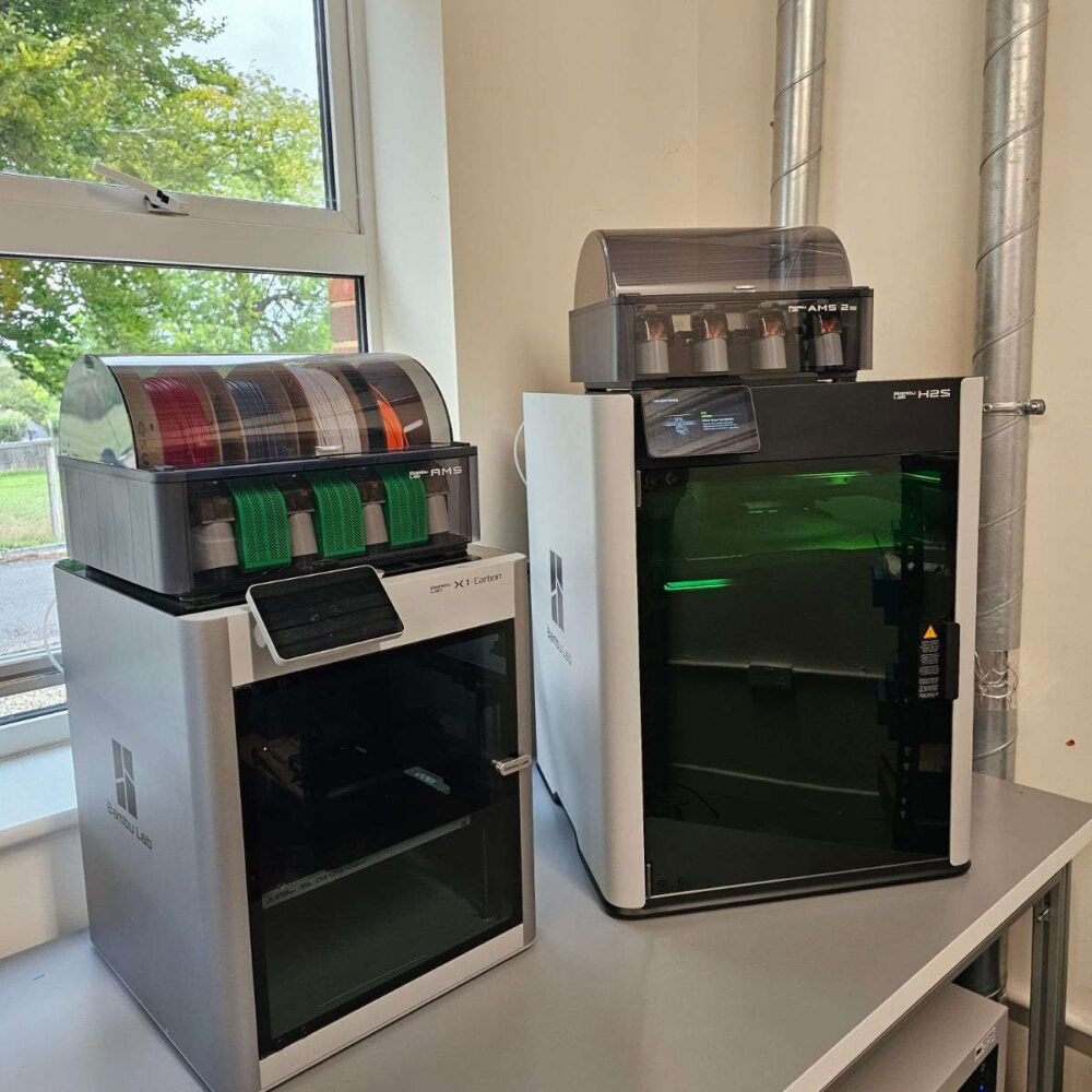 Two 3D printers side by side in an engineering production lab, showing the coloured printer filament reels and the large print bed size of the pH2S printer on the right.